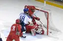 Ice hockey players crowd around a red net as the goalie dives across the crease