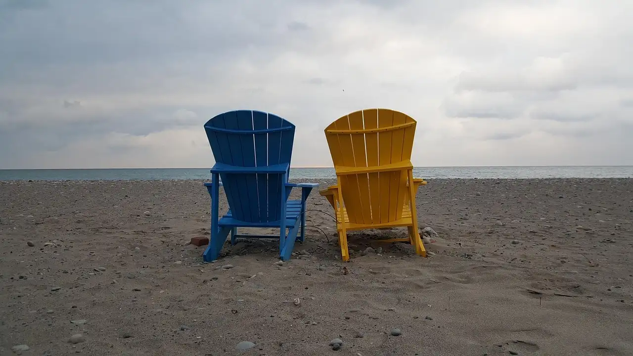 A blue and a yellow Muskoka chair side by side on a pebble beach by Lake Ontario under a cloudy sky.