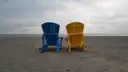 A blue and a yellow Muskoka chair side by side on a pebble beach by Lake Ontario under a cloudy sky.