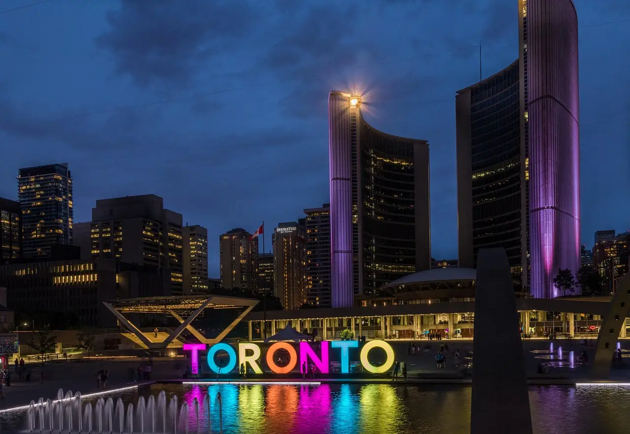 Toronto City Hall's curved towers lit purple above the colourful Toronto sign and the reflecting pool at Nathan Phillips Square at dusk.