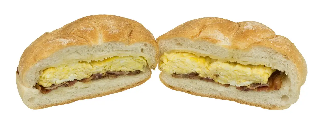 A breakfast sandwich with egg and bacon on a soft bun, cut in half and shown on a white background.
