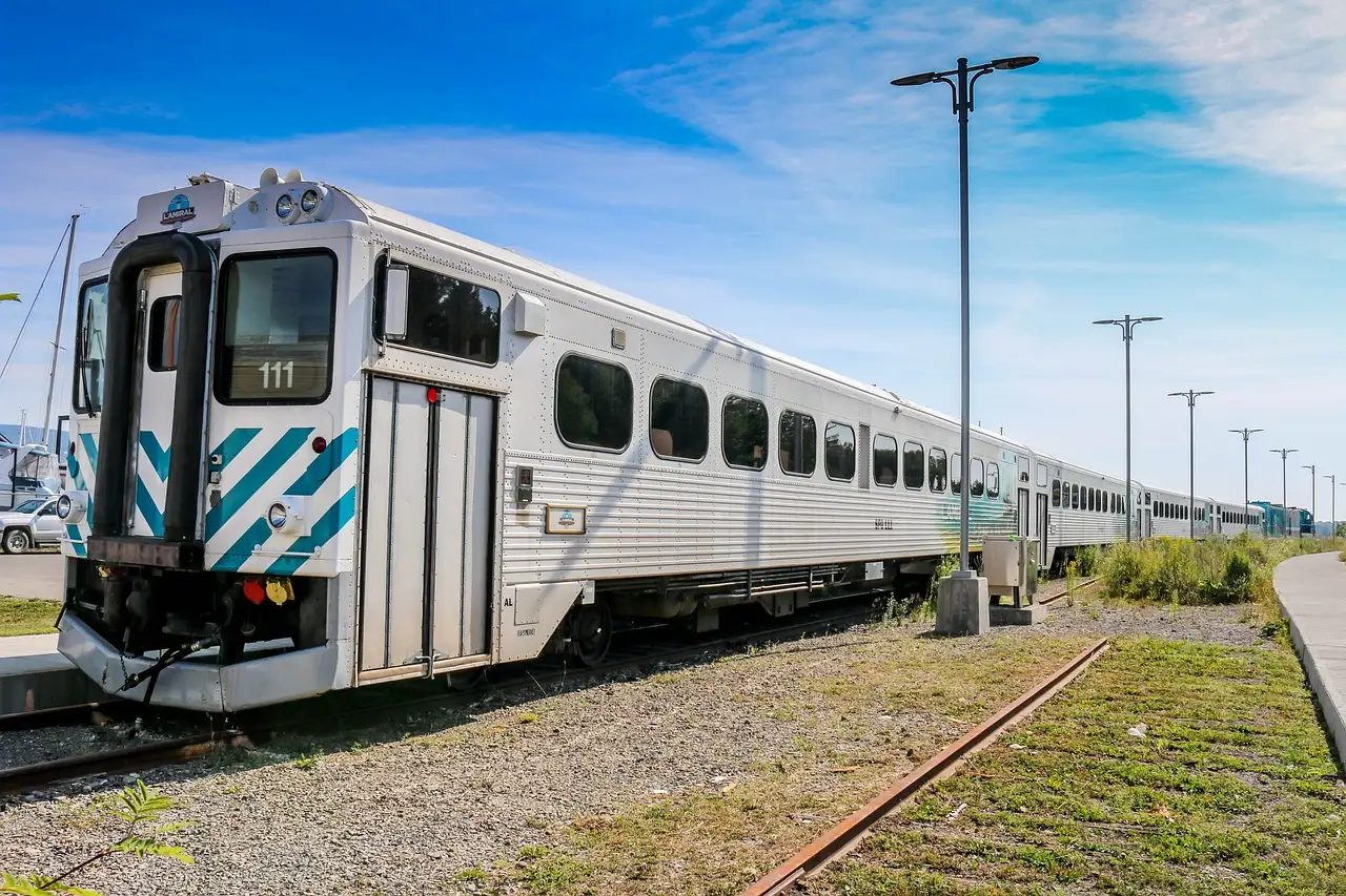 A silver passenger train with teal stripes on its front car standing on a track in summer sun.